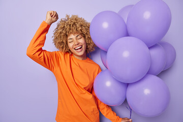 Happy carefree woman with curl hair dances with rhythm of music celebrates special occasion holds bunch of helium balloons enjoys party time wears orange jumper isolated over purple background