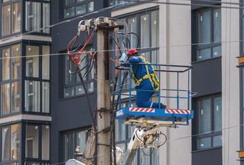 An electrician raised in the cradle of a tower lift mounts wires on a pole against the background of a high-rise building