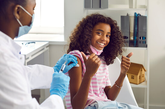 Happy beautiful Afro American school child girl in medical office at clinic looks at camera, smiles and shows thumbs up as she gets arm vaccine injection for good virus immunity. Vaccination concept