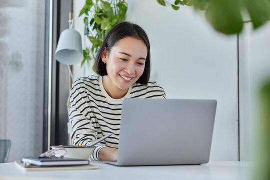 Female remore worker keyboards on laptop computer works from home on startup project smiles happily dressed in striped jumper poses against cozy interior. Asian colleges student studies distantly