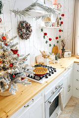 Wooden kitchen worktop with built-in oven and gas hob with a Christmas cake on it. Scandinavian kitchen with a Christmas tree with garland lights and red toys and a cone wreath on the wall