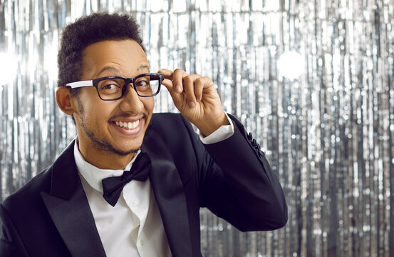 Happy charismatic handsome black guy in modern tux suit, glasses and bow tie having fun at party. Cheerful elegant man on shiny background looking at camera and smiling with broad white toothy smile