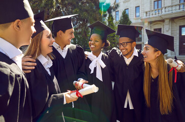 Happy multiracial university graduates walking, talking, laughing and hugging together outdoors. Joyful young male and female students in black robes and academic caps celebrating their graduation.