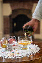 Pouring of apple cider drink in glasses in old french house with fireplace on background, Normandy, France
