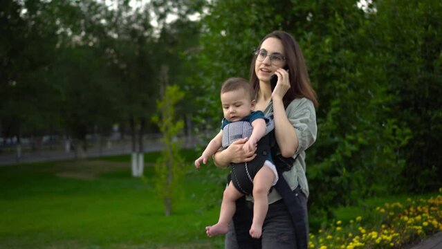 A young mother walks in the park with a child and speaks on the phone. Woman with glasses with her son. Newborn baby in a kangaroo-backpack. Close-up.