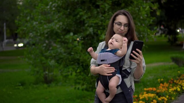 A young woman walks through the park with a child and a phone in her hands. Mom in glasses with her son in her arms. Newborn son in a kangaroo-backpack.