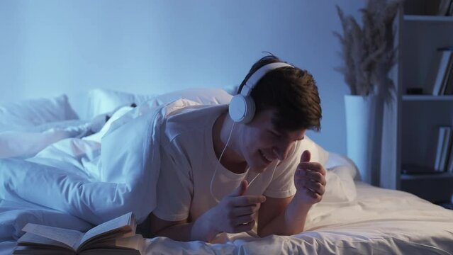 Music in bed. Carefree home evening. Positive playlist. Happy inspired relaxed guy dancing enjoying rythm in headphones in dark bedroom.