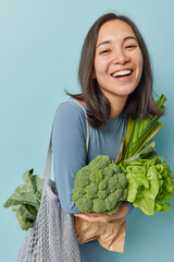 Positive carefree young Asian woman with dark hair smiles broadly shows white teeth poses with fresh green vegetables keeps to healthy nutrition eats organic food isolated over blue background