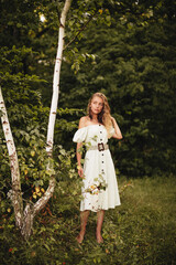 Stylish blonde girl with long curly hair in a light long dress and with earrings with feathers in the summer forest. 