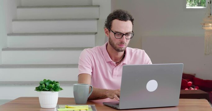Handsome successful young Caucasian man wearing a light pink polo shirt, freelance worker, experienced programmer writing computer programs while working remotely on laptop in comfortable home office.