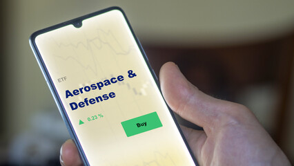 An investor's analyzing the aerospace & defense etf fund on screen. A phone shows the ETF's prices aerospace and defense to invest