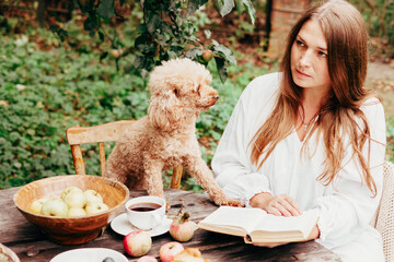 young woman model plus size in white dress sit at table with pet dog poodle, resting and drinking tea after harvesting autumn in back yard, fall and harvest, zero waste life, eco-friendly simple life
