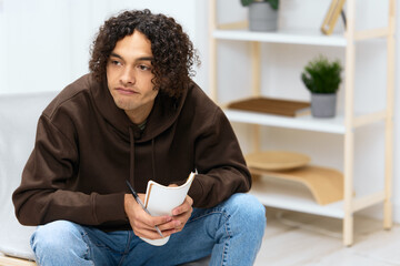 guy with curly hair writes in a notebook sitting on a chair in the room light background