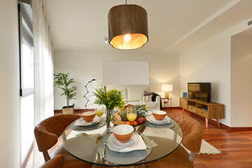 Dining room with round glass table with breakfast service set up with a tray of assorted fruit, vase with flowers, juices, brown upholstered chairs and sideboard with TV