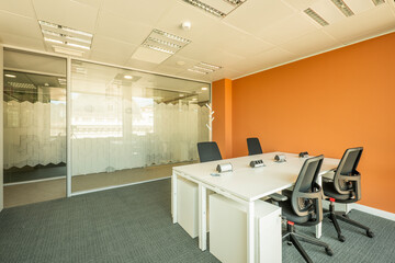 Office cubicle with white furniture, black swivel chairs, sockets on tables, gray carpet floor, plaster ceiling and glass panel as wall