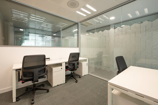 Office cubicle with white wooden tables, black swivel chairs, wall and sliding glass door