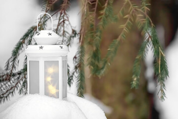 A white lantern on the snow in the forest in winter. Blurry background, light. Space for text. Concept. Festive festival yard decoration for Christmas. Lantern on a winter background with fir branches