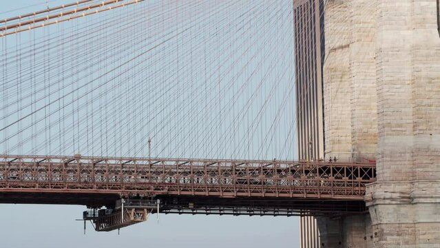 Close view of Brooklyn bridge across the East River. View of cars driving along it and pedestrians walking along it. Ropes and a huge pillar. NYC, USA