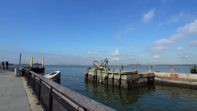 Pier A in sunny day and American Merchant Mariners Memorial: Three figures made of bronze on boat, one figure is trying to grasp another figure that is in the water by Marisol Escobar. NYC, USA