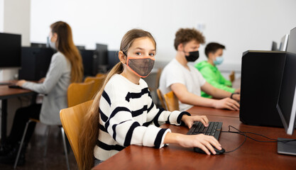 Interested teen girl in protective face mask studying with classmates in school computer class. Necessary pandemic precautions..