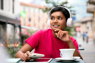 Beautiful smiling asian woman listening music wearing wireless headphones, drinking coffee in cafe, looking away. Coffee break concept 