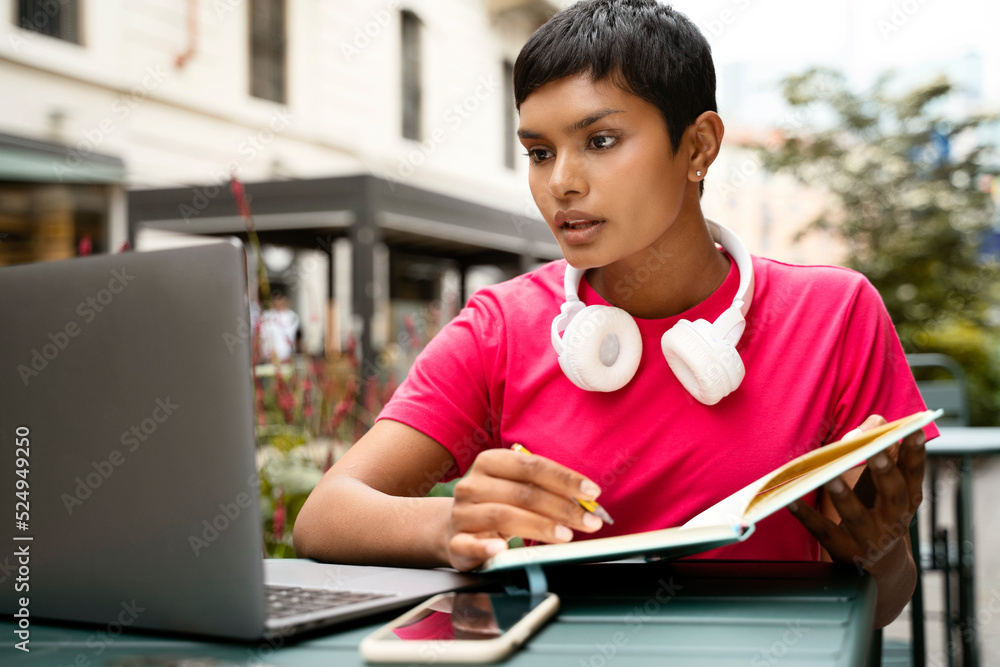 Wall mural Beautiful pensive Indian student using laptop computer studying, taking notes, online education concept. Freelancer working online sitting at workplace. Asian woman watching training courses 