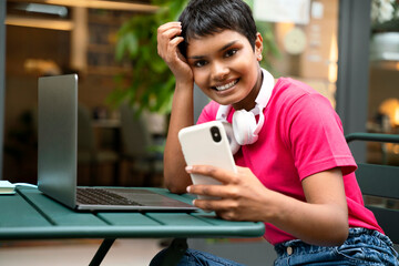 Portrait of smiling asian freelancer using laptop, holding mobile phone working online sitting at workplace. Happy successful Indian woman using mobile app shopping online looking at camera 