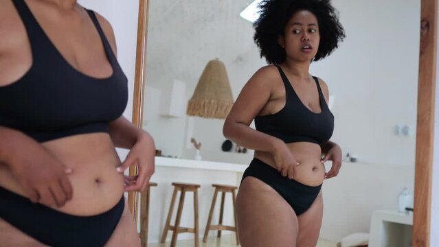 An overweight African American woman looks at herself in the mirror, draws in a fat belly, a woman has stress due to being overweight. American women are prone to obesity due to eating fast food.