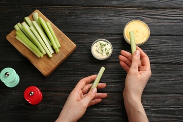 Woman dipping celery stick in sauce at dark wooden table, top view