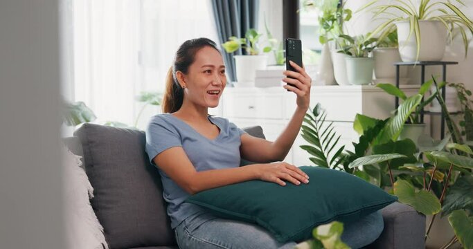 Young Asian woman wear casual site on couch use smartphone raise hand video call to say hello and talk to family and friend with happiness and smiling in living room indoor plants at home.