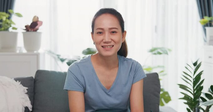 Young Asian woman wear casual site on couch looking at camera raise hand video call to say hello and talk to family and friend with happiness and smiling in living room indoor plants at home.