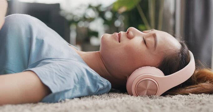 Young Asian university woman with casual wear headphone lay down on carpet floor listen music calm and relax take a rest in living room at home. Cozy home interior with indoor plants concept.