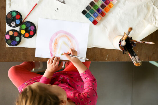 top shot of a girl wearing pink painting rainbow with water colour, paper and paint brushes