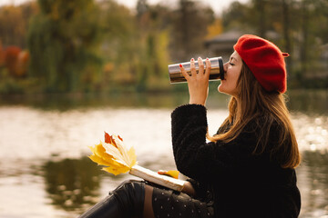 Woman in a red beret reading book on wooden pontoon. Autumn season. Female with trendy clothes...