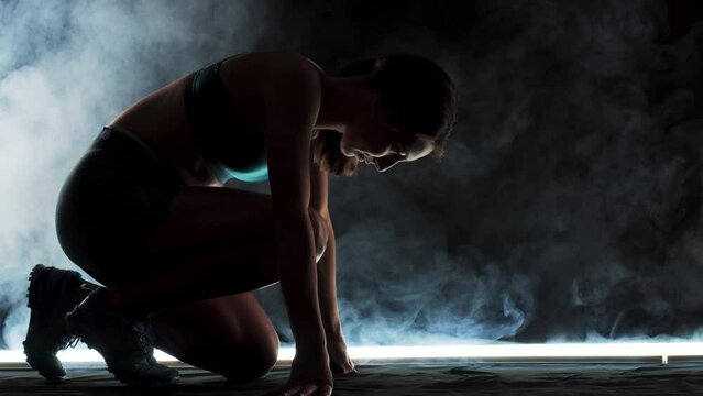 Silhouette female runner in race  start position. Girl in sportswear posing on lit track with smoke in the background.