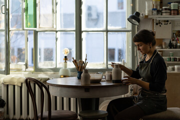 Woman concentrated on modeling jug from raw clay enjoys painting vase. Lady sits at table smiling against window. Female ceramic business owner makes craft for sale in handmade potter retail store
