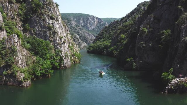 Small boat with tourists sails between the steep mountain sides over Lake Matka in the major tourist attraction Matka Gorge of northern marcedonia on a sunny summer day. Drone dolley shot