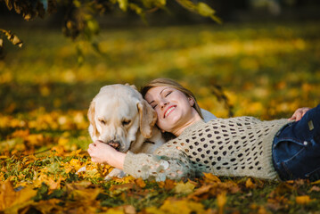 A beautiful blonde lies in yellow leaves in a park and plays with her labrador.