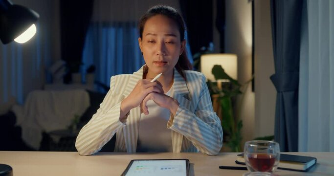 Young happy Asian businesswoman wear formal sit in front of desk with tablet hold stylus looking at camera video online meeting with colleague in cozy living room at home night. Overtime work concept.