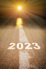 New year 2023 on road surface with white marking line leading into abstract blur sunlight and sun. start sustainable lifestyle concept and motivation success idea