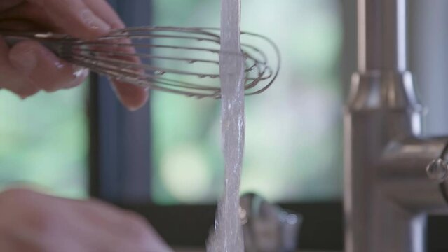 A Caucasian male is cleaning and rinsing a French whisk in a home kitchen. Close-up shot. Slow motion.