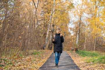 A teenage boy walks through an autumn park and shoots a video. A Caucasian boy in a black jacket holds a monopod with a smartphone. Blogger. Emotional communications..