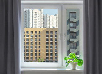 View through the window on facades of buildings. Dense urban development background. Interior space with dark grey curtains and indoor plant.