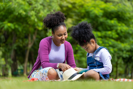 African American mother is teaching her young daughter to read while having a summer picnic in the public park for education and happiness concept