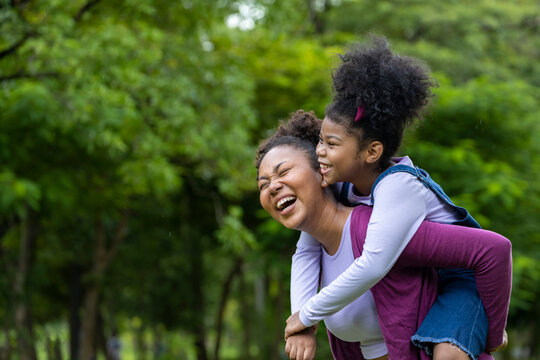 African American mother is playing piggyback riding with her young daughter while having a summer picnic in the public park for wellbeing and happiness concept with copy space