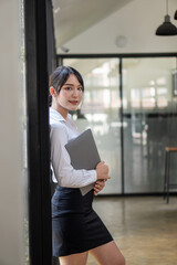 Fototapeta premium A young accountant stands with her laptop next to a glass wall in her office.