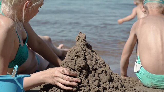 The mother of two sons came to the beach on this hot summer day. Children are busy with their own interests. One builds sand figures, the other splashes in the water. The woman helps them.