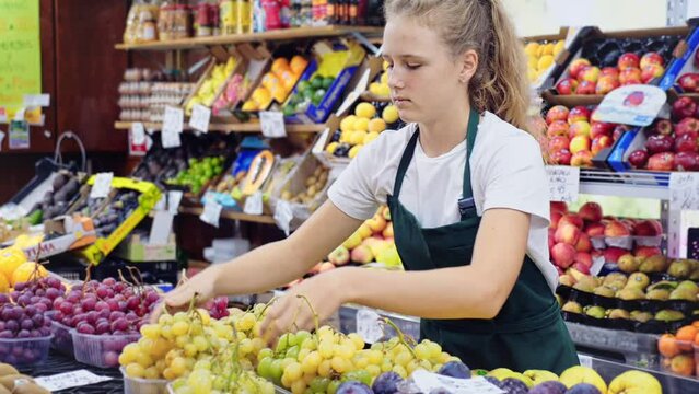 Portrait of a smiling fifteen-year-old girl who works part-time in a store as a trainee seller, standing at the counter, holding a box of grapes in her hands. High quality FullHD footage