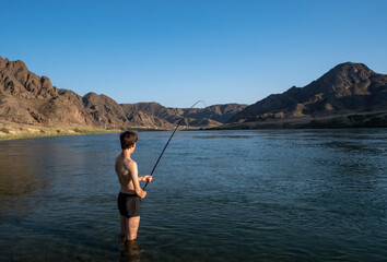 A young guy is fishing in the river with a float rod. The guy is standing in the water with a fishing rod.