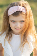 Vertical portrait of a child of model appearance. A girl with blonde hair and blue eyes in a white dress. A child with a pink headband. Beautiful blue eyes
